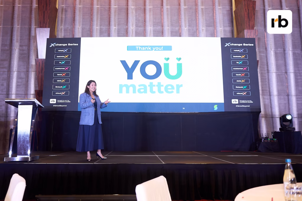 Woman in a blue suit speaks on stage. Behind her, a large screen displays "YOU matter" and "Thank you!" The mood is positive and motivational.