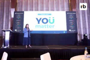 Woman in a blue suit speaks on stage. Behind her, a large screen displays "YOU matter" and "Thank you!" The mood is positive and motivational.